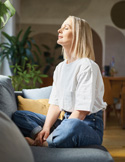 A woman sitting on a couch with her eyes closed, practicing mindfulness in a bright, peaceful living room.