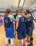 A team of senior women basketball players wearing blue uniforms huddling together on a court.