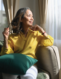 An older woman in a yellow sweater sits by a window, smiling thoughtfully as sunlight fills the room.