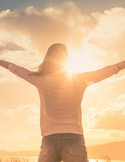 Silhouette of a woman with arms outstretched toward a bright, golden sunset over a scenic landscape.