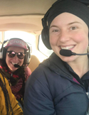Two smiling women wearing aviation headsets inside a small airplane cockpit, looking at the camera.