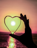 A hand holds a heart-shaped glass against a sunset over the ocean, with the sun centered inside the heart.