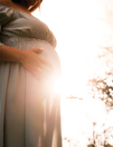A pregnant woman stands outdoors, her hands resting on her belly with bright sunlight creating a warm glow.