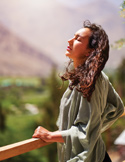 A woman with her eyes closed tilts her head back toward the sun, standing on a balcony with mountains behind her.