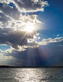 Sunlight breaking through dramatic clouds over a calm body of water, with bright rays streaming toward the horizon.
