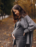 Woman standing on a leafy path in autumn, smiling and gently holding her belly while wearing a gray dress and patterned coat.