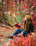 Woman sitting among vibrant autumn leaves, holding an open book and looking upward while surrounded by red and orange foliage.
