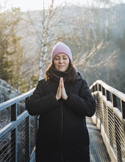 Woman standing on an outdoor walkway in winter, wearing a coat and knit hat, with hands together in a peaceful, prayer-like pose.
