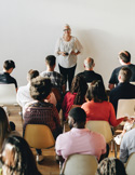 Speaker standing at the front of a room addressing a seated audience during a class or workshop.