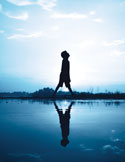 Silhouette of a person walking across a still body of water, reflected perfectly against a bright, blue sky.