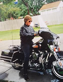 Woman in a black leather jacket and sunglasses sitting on a black cruiser motorcycle in front of a wooden fence.