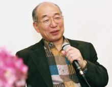 Hiroshi Doi Sensei Hiroshi Doi Sensei wearing glasses and a patterned shirt is smiling and holding a microphone, seated against a plain background.