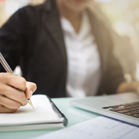 Person in business attire writing in a notebook beside an open laptop on a bright desk.