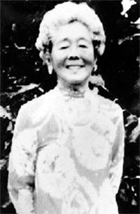Black-and-white photo of Hawayo Takata smiling, wearing a patterned dress, standing outdoors in front of dark foliage.