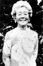 Black-and-white photo of Hawayo Takata smiling, wearing a patterned dress, standing outdoors in front of dark foliage.