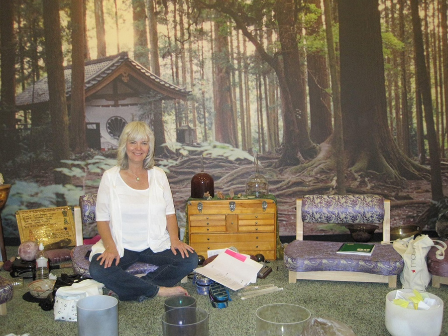 Laurelle Gaia sits cross-legged indoors, surrounded by objects, with a forest and cottage mural in the background.