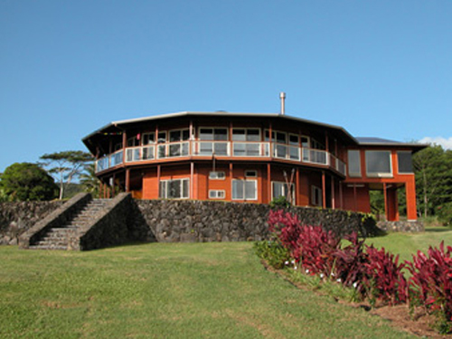 Round, two-story building with large windows and a stone base, set on a grassy. landscaped hill.