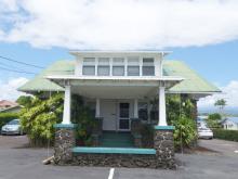 Small gray three-story building with narrow windows and rooftop units beside a fenced, mostly empty parking lot on a quiet street.
