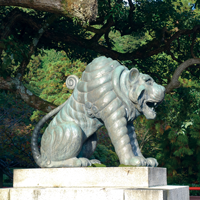 Bronze tiger statue with a stylized, ridged mane, perched on a stone platform against a backdrop of green trees located at the Main Temple on Kurama Yama, Japan.