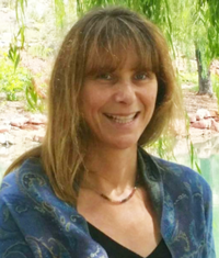 Smiling woman with straight brown hair, wearing a blue patterned shawl, outdoors by greenery.